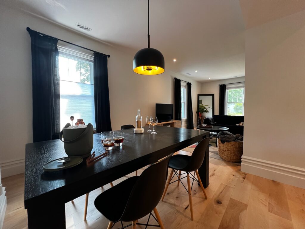 Photo of the modern dining area at SquirrelHaus PEC. Tall windows with dark curtains, black table & chairs, bright wood floors.