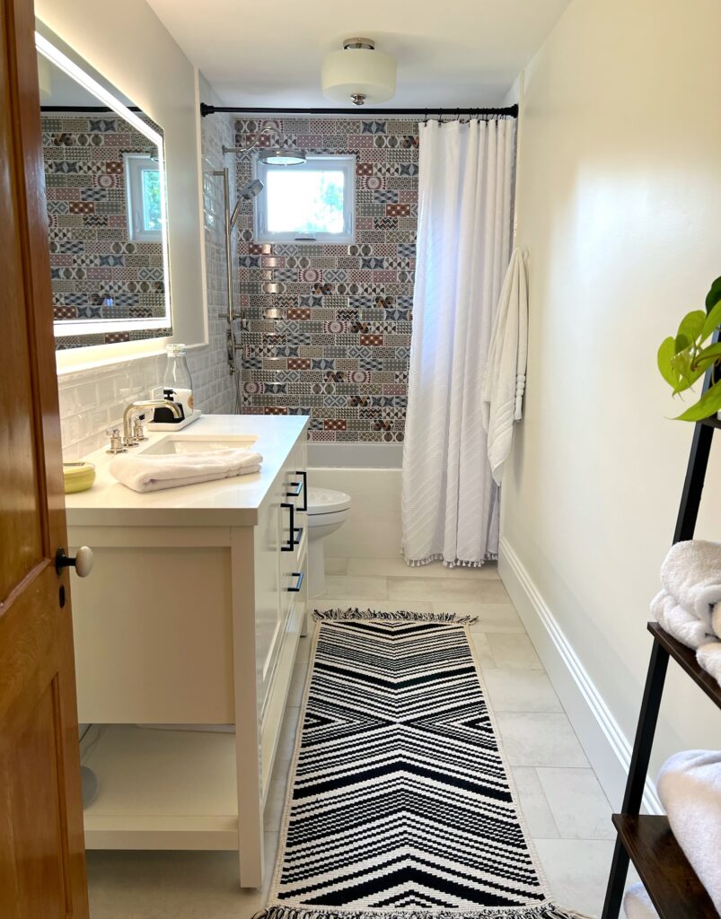 SquirrelHaus PEC's long, narrow bathroom with a small window in the bathing area at the end, a chevron rug, wooden door, and a white vanity with a light-mirror above