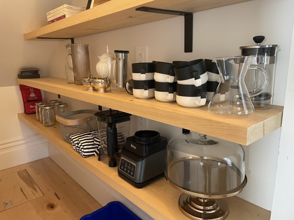 Photo of the pantry at SquirrelHaus PEC - open shelving stocked with mugs, carafes, a covered cake server, aprons, cookbooks, pitchers, a teapot and more.