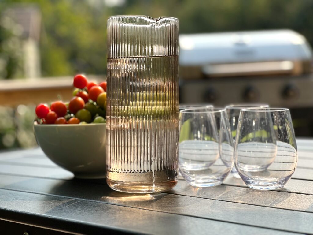 A ribbed pitcher of water, bowl of cherry tomatoes, and glasses on a black outdoor dining table at SquirrelHaus PEC. A grill is in the background.