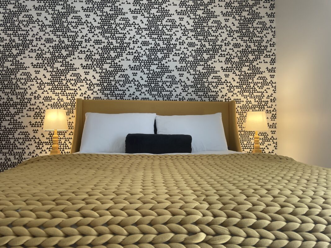 Photo of a yellow upholstered bed with a chunky knitted blanket, pillows, a black velvet bolster and two lit yellow bedside lamps at SquirrelHaus PEC