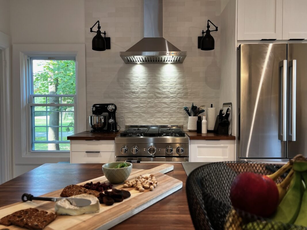 Photo across the island of SquirrelHaus PEC's modern kitchen with commercial-style appliances and tall window letting in natural light
