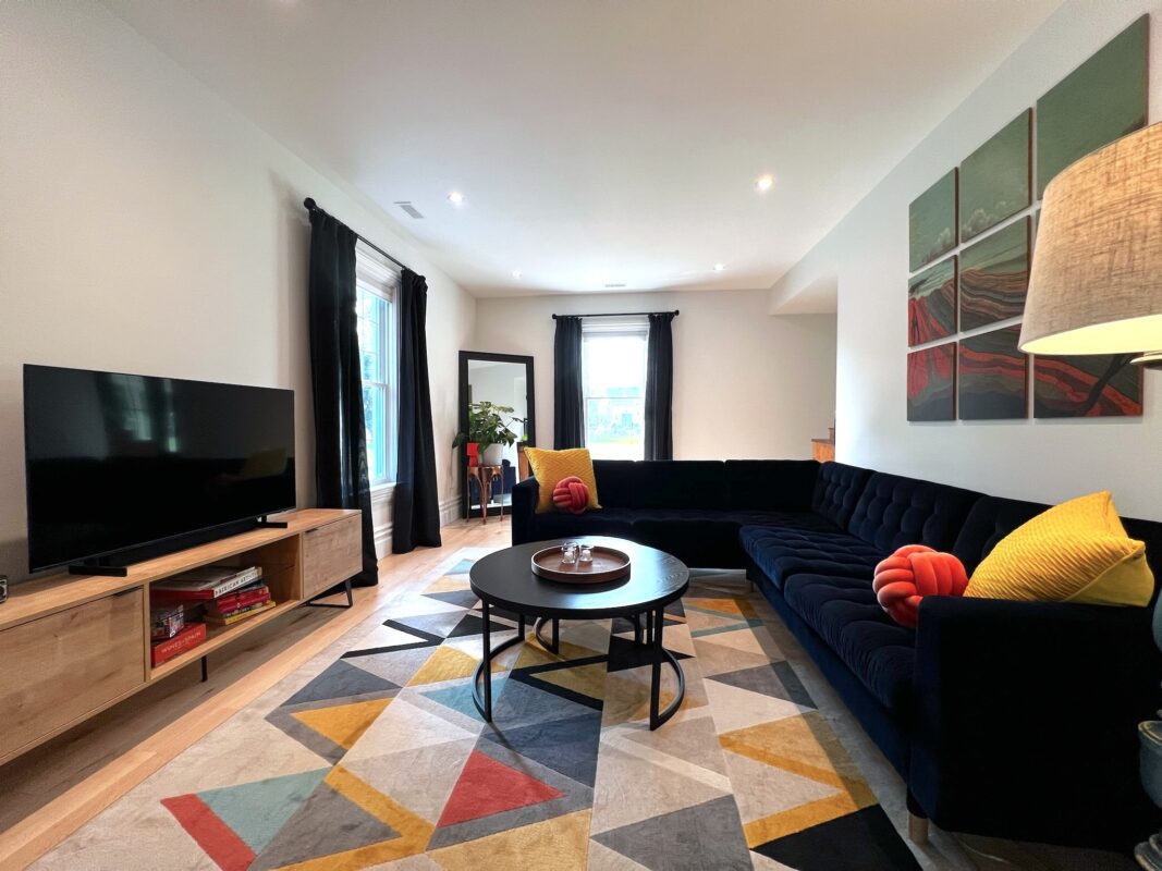 SquirrelHaus PEC's main living space with a velvet sectional, graphic colourful rug, nested round coffee tables, a large entertainment centre, and tall windows filling the space with natural light.