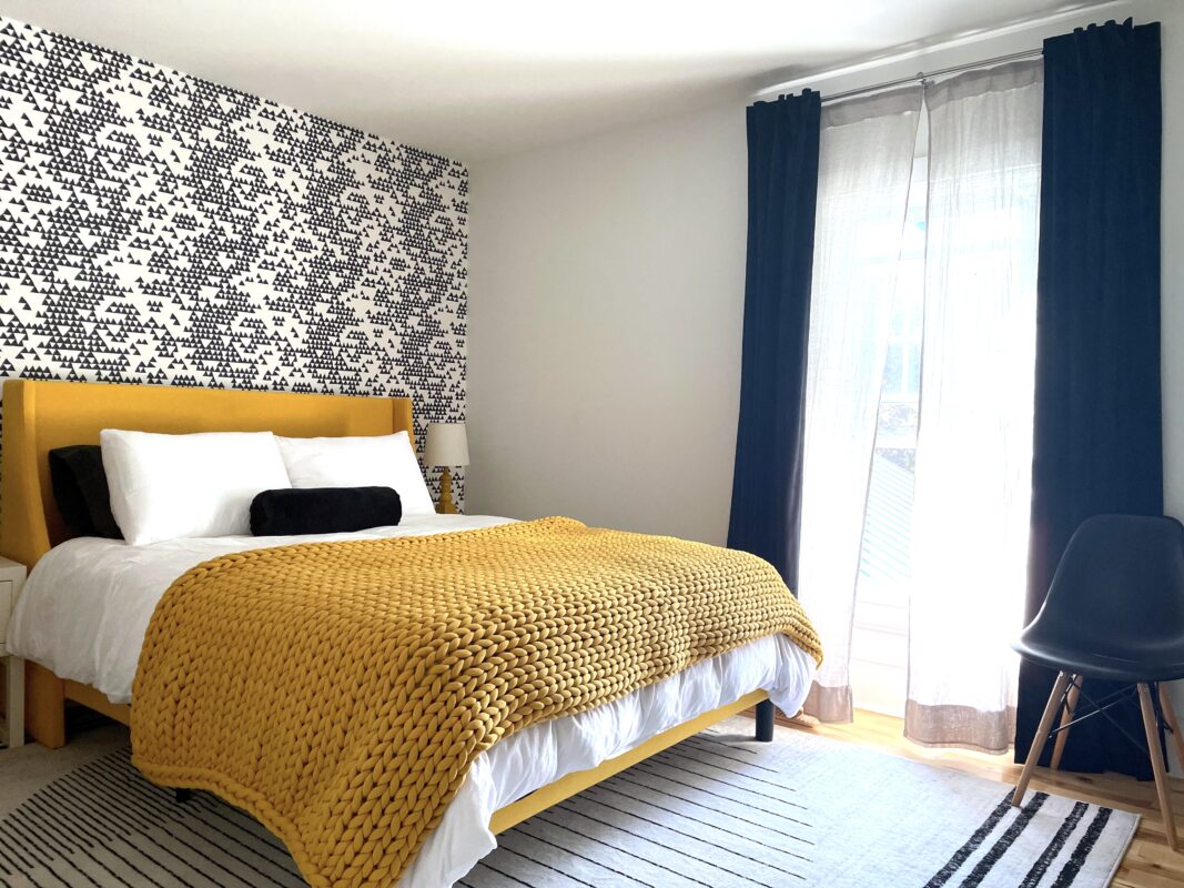 SquirrelHaus PEC's warm middle bedroom featuring a yellow upholstered queen bed, graphic rug, black and white modern wallpaper, black velvet drapery and a chunky knit yellow weighted blanket.