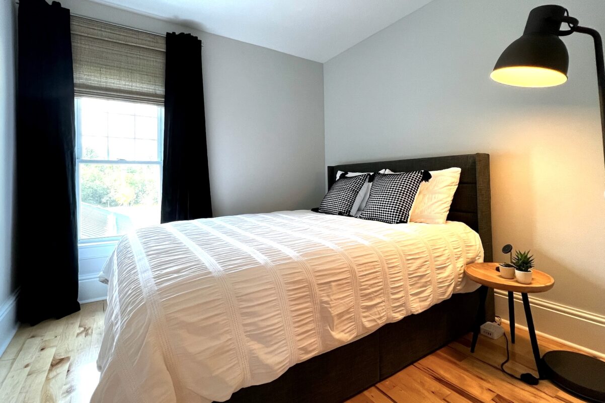 Bedroom at SquirrelHaus PEC with a linen-upholstered bed, crisp white duvet, dark velvet curtains, a night table and standing lamp on wood flooring